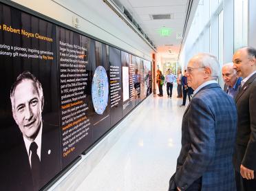Three people look at a TV screen displaying information about Robert Noyce